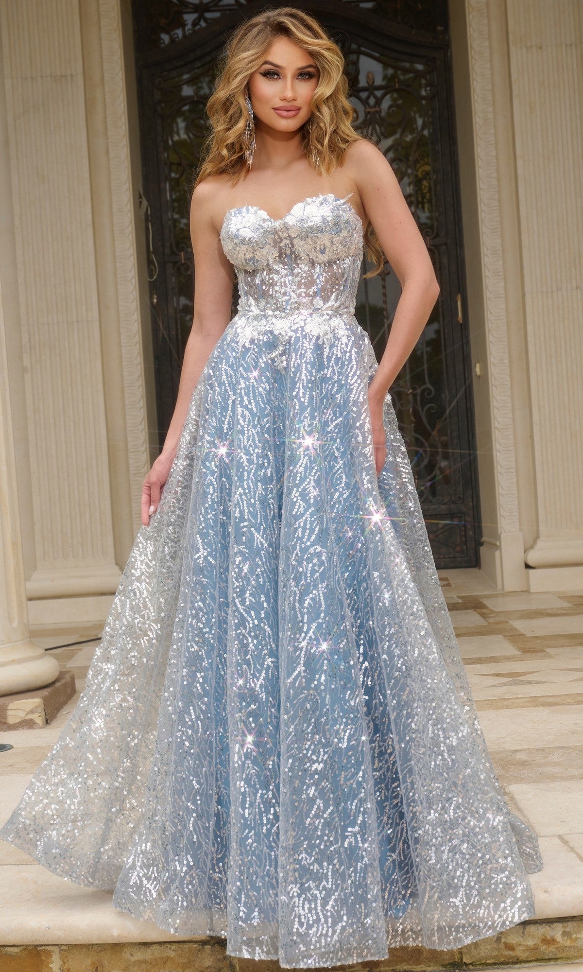 A woman stands on stone steps before double doors, wearing the Jovani 08417—a strapless long sequin prom ball gown in silver and blue with a sweetheart neckline and full skirt, perfect for making an entrance.